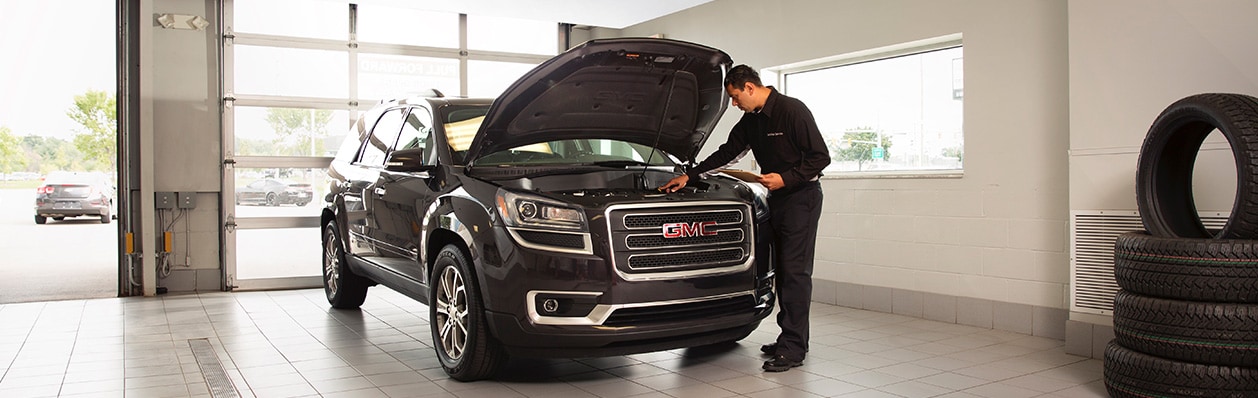 A service technician dressed in black looks under the hood of a black GMC Acadia inside a bright and clean garage